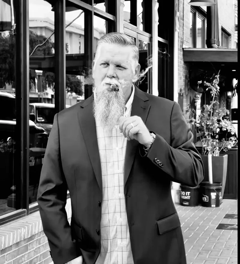 Man with beard smoking cigar outside building.